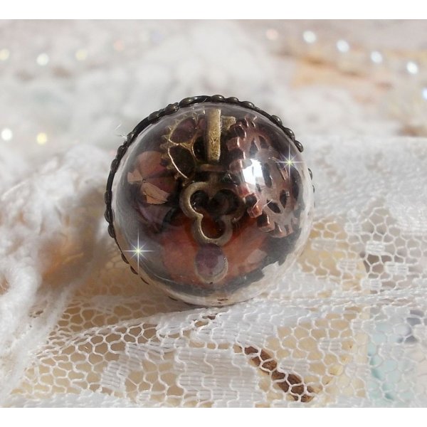 Bague Majest&eacute; Steampunk aux rouages m&eacute;caniques &agrave; laquelle s&rsquo;ajoute une cl&eacute; et un strass de couleur Siam et un strass en verre Dor&eacute;. Le tout est mis sous un globe en verre sur une bague r&eacute;glable laiton