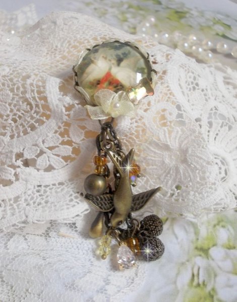 Broche Colombes aux plumages blancs avec des breloques filigran&eacute;es gouttes et lisses, perles rondes et n&oelig;ud en cristal, ruban organza cr&egrave;me, pendentif plaqu&eacute; or avec un zircon sur une broche en m&eacute;tal