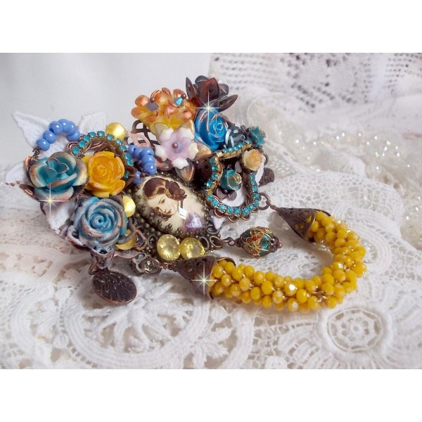 Broche Eclats P&eacute;tillants d&rsquo;inspiration Baroque, orn&eacute;e de fleurs en r&eacute;sine autour d&rsquo;un cabochon strass&eacute;s et perl&eacute;s sur estampes, cristaux, breloques, perle cloisonn&eacute;e, perles jaunes en verre et roses. 