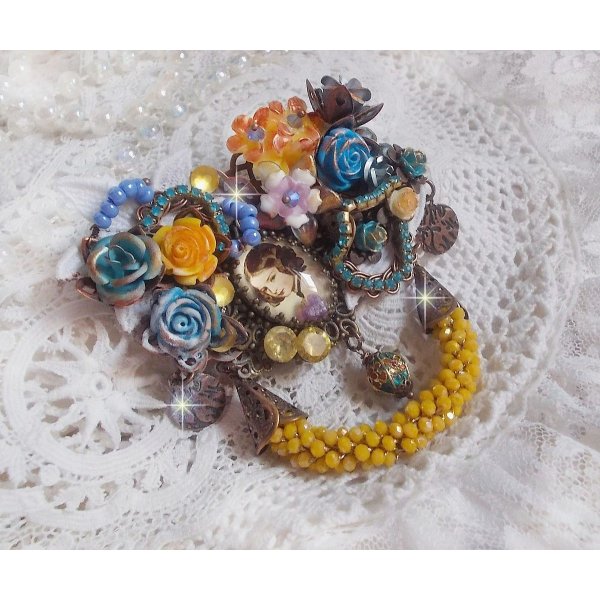 Broche Eclats P&eacute;tillants d&rsquo;inspiration Baroque, orn&eacute;e de fleurs en r&eacute;sine autour d&rsquo;un cabochon strass&eacute;s et perl&eacute;s sur estampes, cristaux, breloques, perle cloisonn&eacute;e, perles jaunes en verre et roses. 